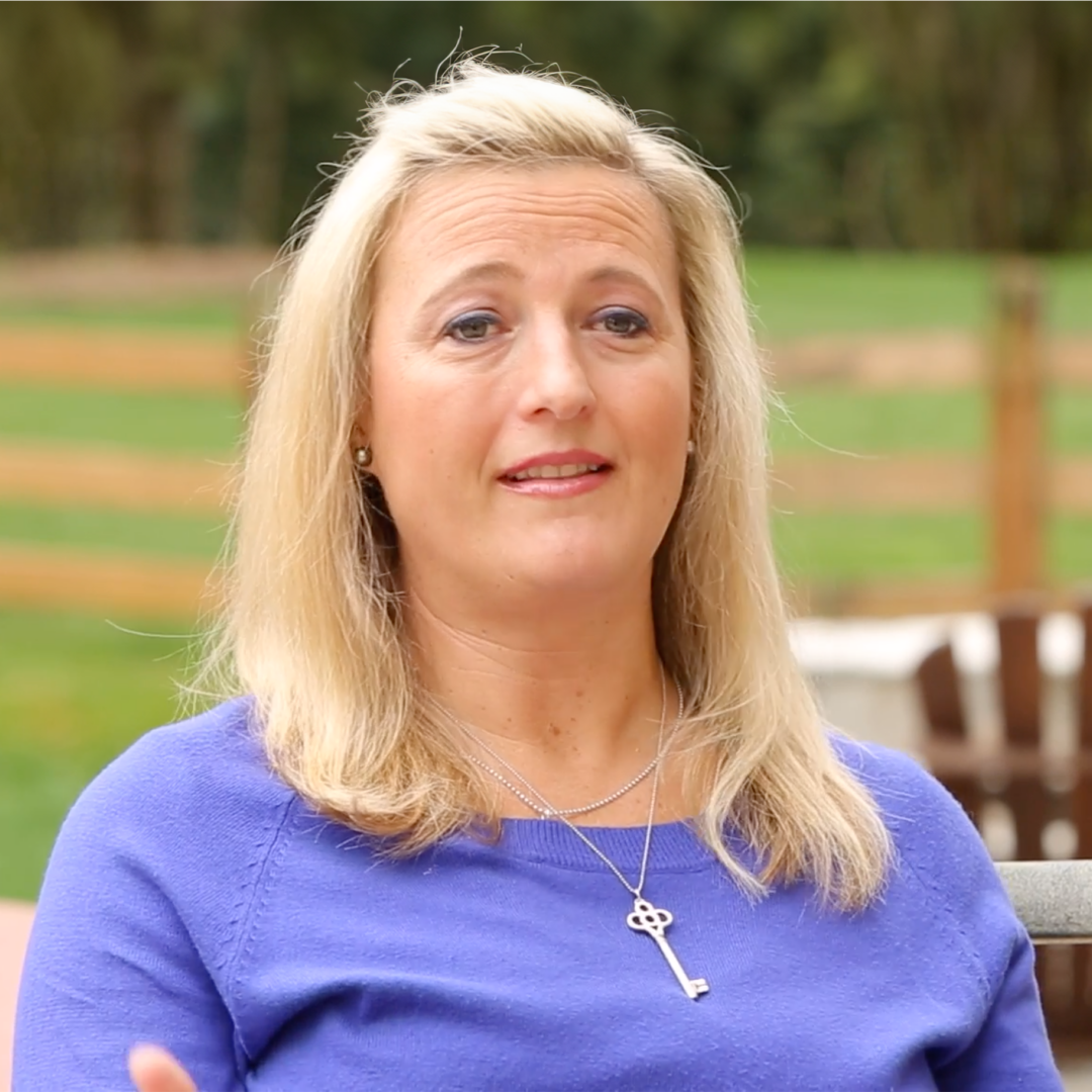 A woman with long blonde hair wearing a purple sweater and a key-shaped necklace sits outdoors, with a blurred green background and a wooden fence behind her.